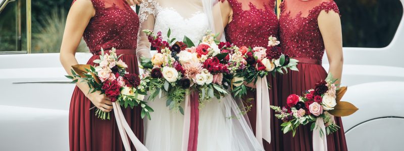 bride in white wedding dress with her three bridesmaids in red dresses holding flower bouquet